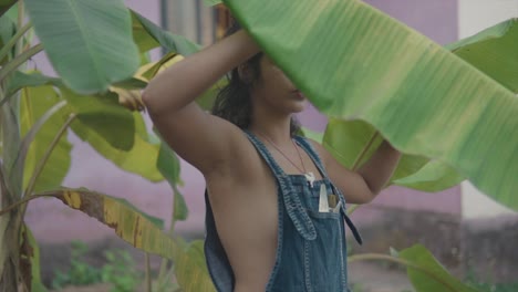 medium handheld shot of a beautiful indian model dressed in a blue jean dress, chain and a tightening hair elastic on her arm lifting a large leaf of a plant in the garden