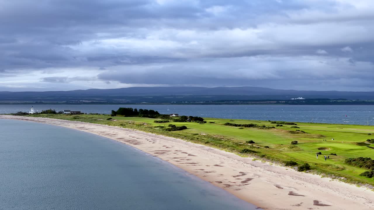 Drone glides above sandy beach, grassy hills, and calm sea under dramatic, cloudy summer sky