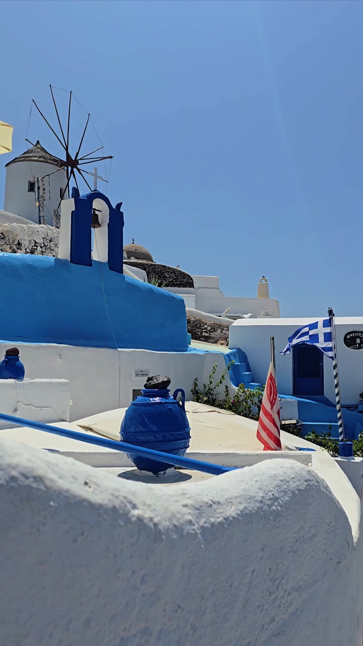 Vista ensolarada de verão de Santorini na Grécia com casas brancas tradicionais, detalhes azuis, praia de areia, Mar Egeu claro e luz mediterrânea brilhante, criando belas imagens de viagem costeira