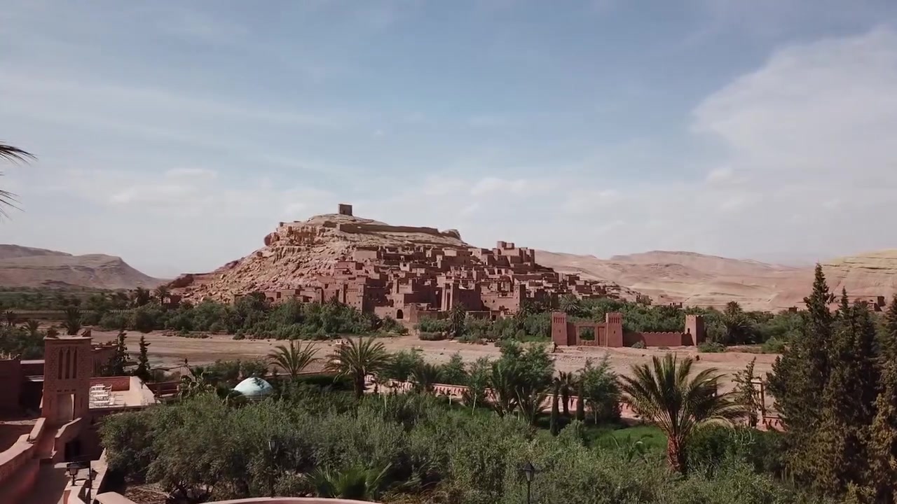 Vista aerea cinematografica della kasbah di Ait Benhaddou che si erge sopra un'oasi lussureggiante con palme a Ouarzazate, Marocco. Iconico sito patrimonio UNESCO circondato da paesaggio desertico, ideale per viaggi, cultura.