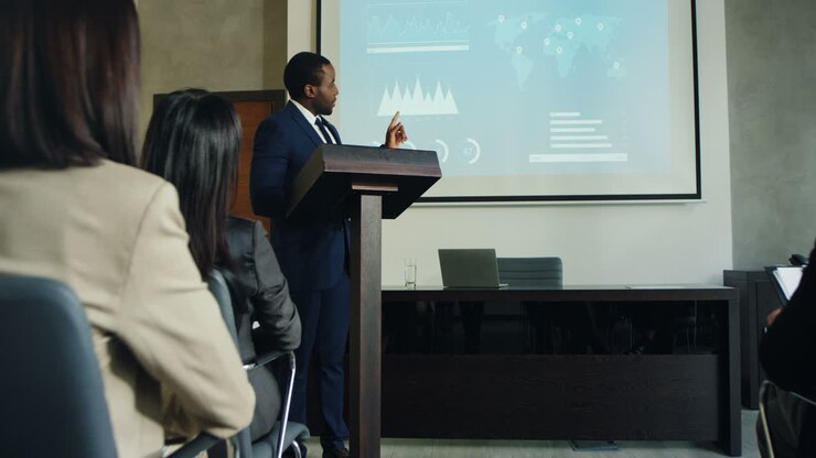 zoom de la caméra sur un homme d'affaires afro-américain parlant sur un podium dans une salle de conférence et montrant des tableaux et des graphiques sur grand écran