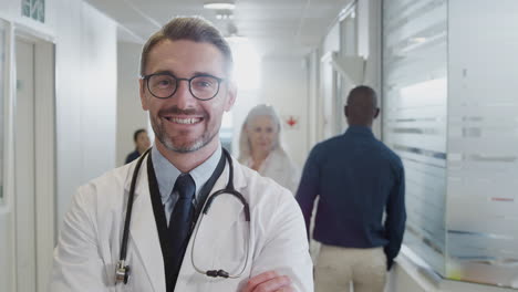 Portrait-Of-Mature-Male-Doctor-Wearing-White-Coat-With-Stethoscope-In-Busy-Hospital-Corridor