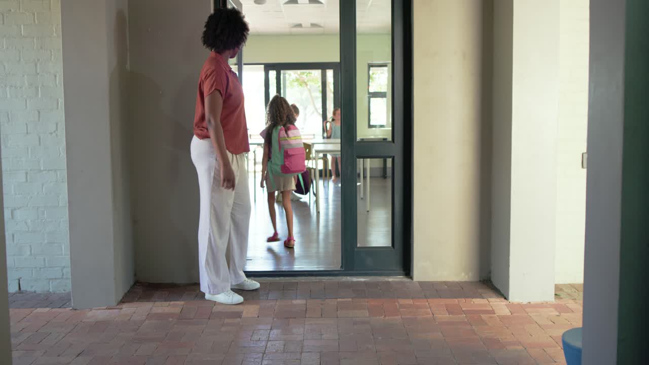 African American teacher welcoming kids entering hall, guiding backpacks through door to classroom
