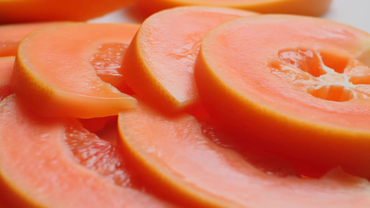 Nærbillede af skåret papaya, der viser levende frugtkød og blank orange tekstur, stablede skiver på hvid baggrund.
