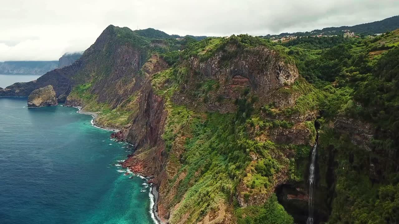 Lush vegetation covers the rocky cliffs of Miradouro da Quinta do Furão as a waterfall cascades into the ocean below, creating a breathtaking scene in Madeira, Portugal