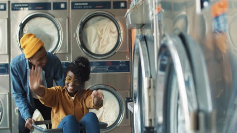 young african american cheerful couple having fun in laundry service room. pretty happy girl sitting in carriage and guy riding her at working washing machines while cleaning clothes.