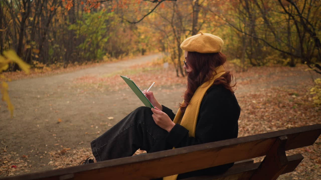 vista lateral de la artista sentada en un bosque sereno, profundamente enfocada en dibujar en su libro, rodeada de colores de otoño y un entorno al aire libre tranquilo
