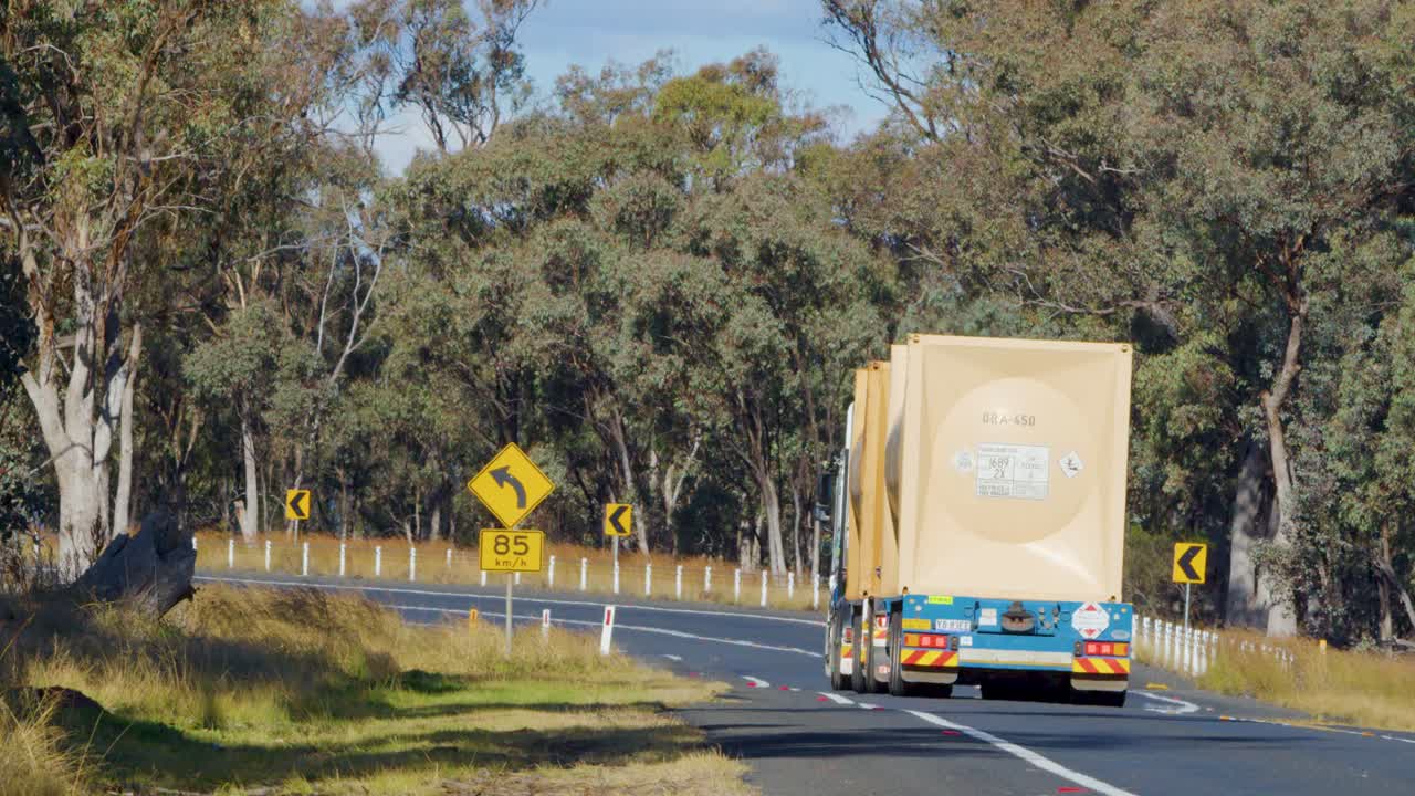 A large road train truck drives along a sunlit, winding rural highway bordered by trees in Coonabarabran, New South Wales, Australia. Static rear view