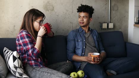 Cute-young-couple,-attractive-caucasian-girl-and-her-handsome-mulatto-boyfriend-sitting-on-the-couch,-drinking-tea,-enjoying-their-time-together.