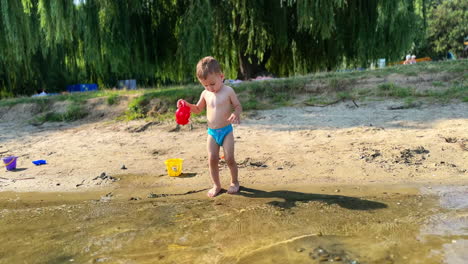 caucasian toddler playing at river bank on hot summer day. cute boy steps slowly into water and fills his red can.