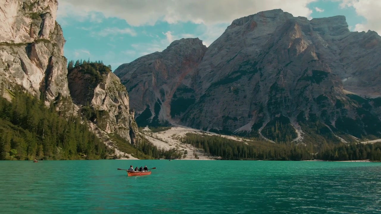 Inquadratura di interesse di una barca con turisti che remano su un lago alpino color turchese, con alte montagne rocciose sullo sfondo, giornata di sole sulle Alpi, cielo azzurro, color grading cinematografico