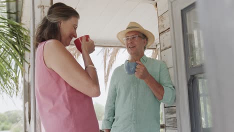 happy senior caucasian couple drinking coffee and talking on porch of beach house, in slow motion