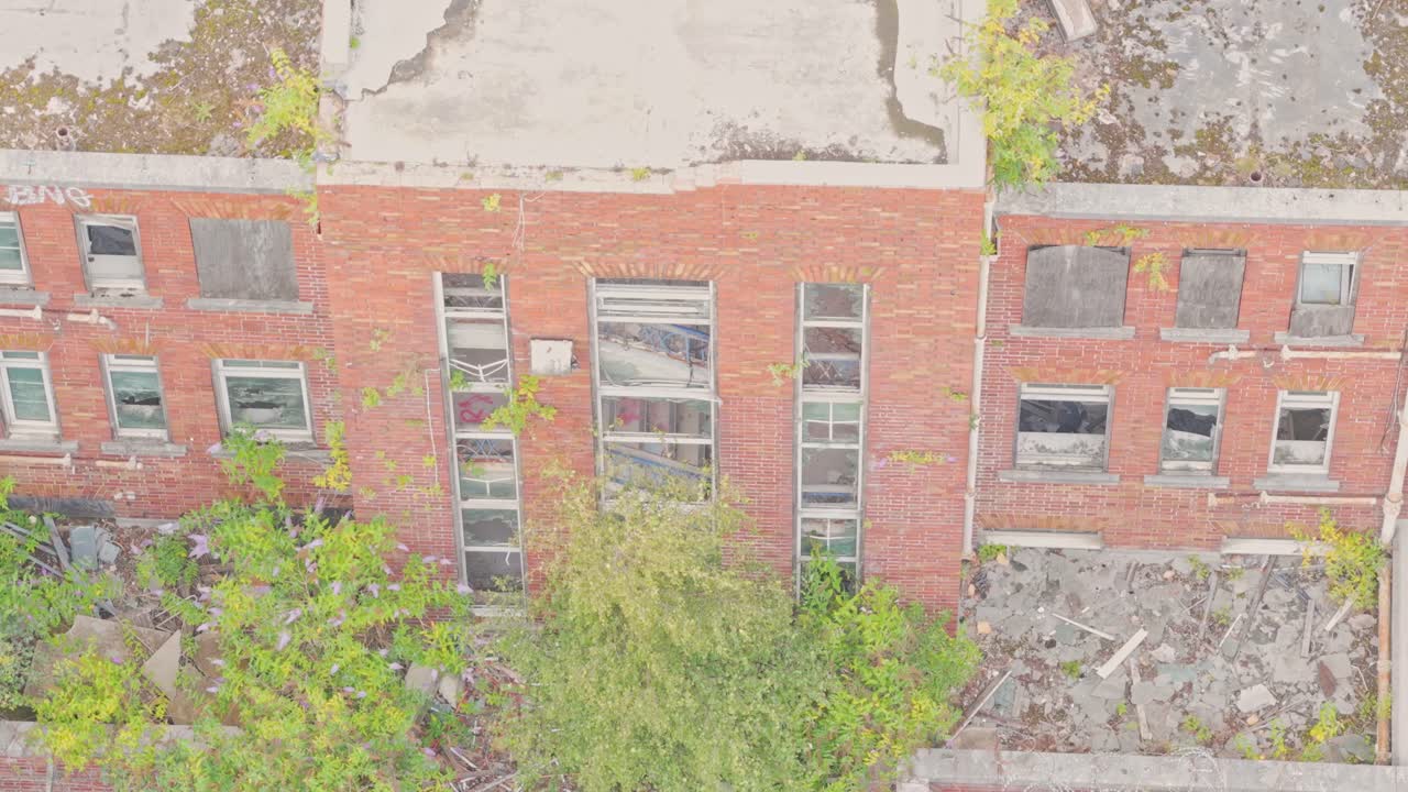 Drone ascends past the facade of the abandoned Salford Crescent Police Station. The red-brick building shows broken windows, boarded openings, and vegetation sprouting from its roof and walls
