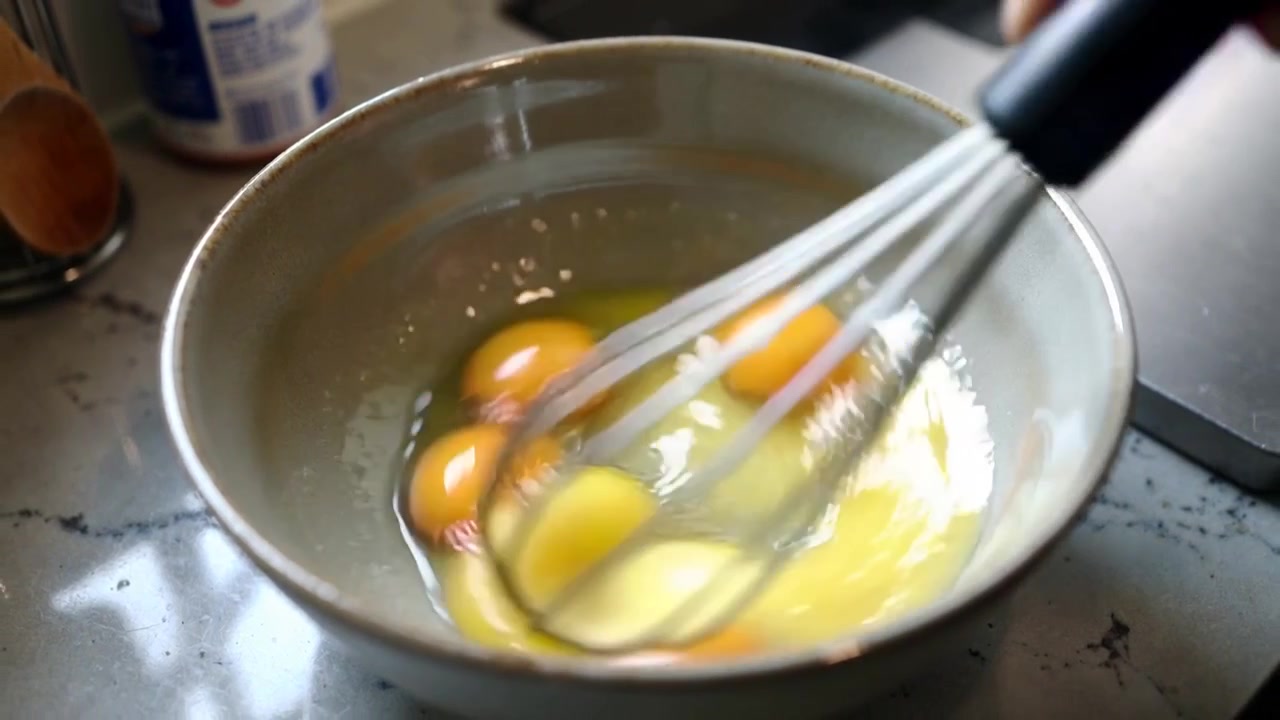 Whisking metal whisk entering breaking four yolks in ceramic bowl at kitchen counter to blend eggs
