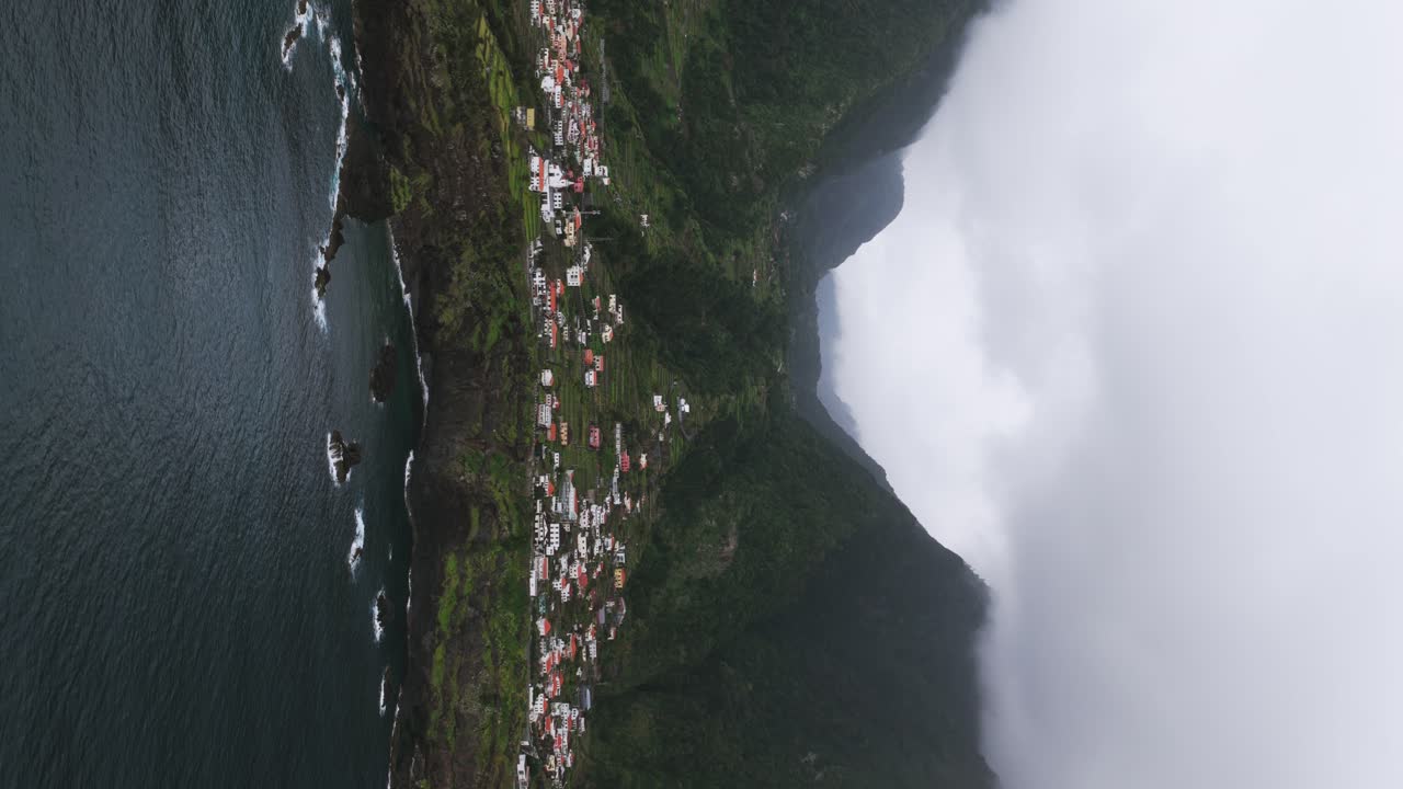 Aerial vertical video view of Seixal village on Madeira coastline meeting rough Atlantic ocean with dramatic sky and mountains