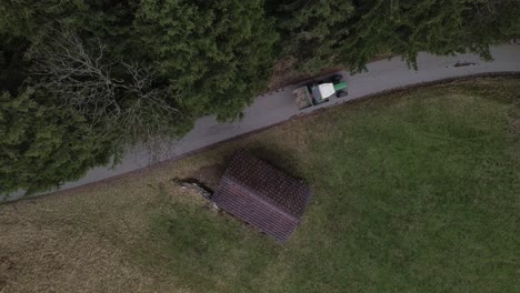 top down aerial drone shot of tractor driving along mountain road with forest and small hut next to the road