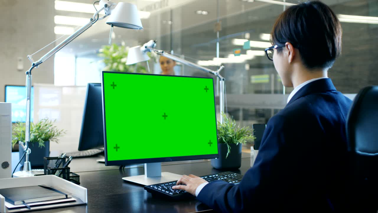 사무실에서 사업가는 모의 녹색 화면을 가진 개인 컴퓨터에서 그의 책상에서 일합니다. 동료는 사무실에 들어와 그녀의 책상에서 일어납니다.