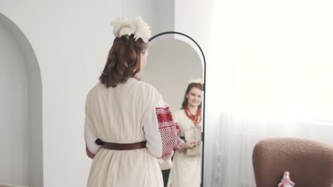 beautiful, charming young woman in embroidered authentic ukrainian attire with flowers in her hair and a handbag poses in front of a mirror with a smile