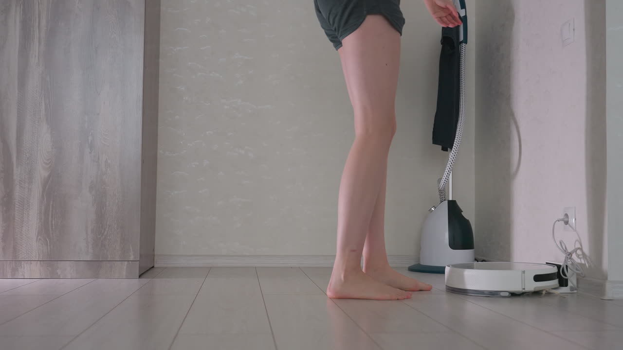 Lady bends to put on item while standing barefoot near robot vacuum charging on wooden floor in modern home interior, natural light highlighting daily routine and technology in domestic environment