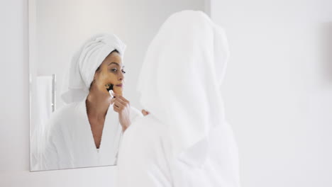 happy biracial woman applying beauty face mask looking in mirror in bathroom, slow motion