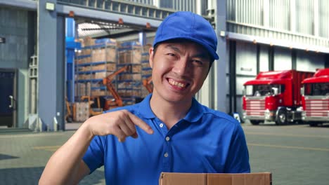 close up of asian male courier in blue uniform smiling and pointing to a carton in hand while delivering it, outside of logistics distributions warehouse