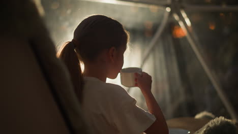 child drinks from cup in morning. girl enjoys warmth of favorite beverage sitting in room against early rays of sunlight. cozy morning at home