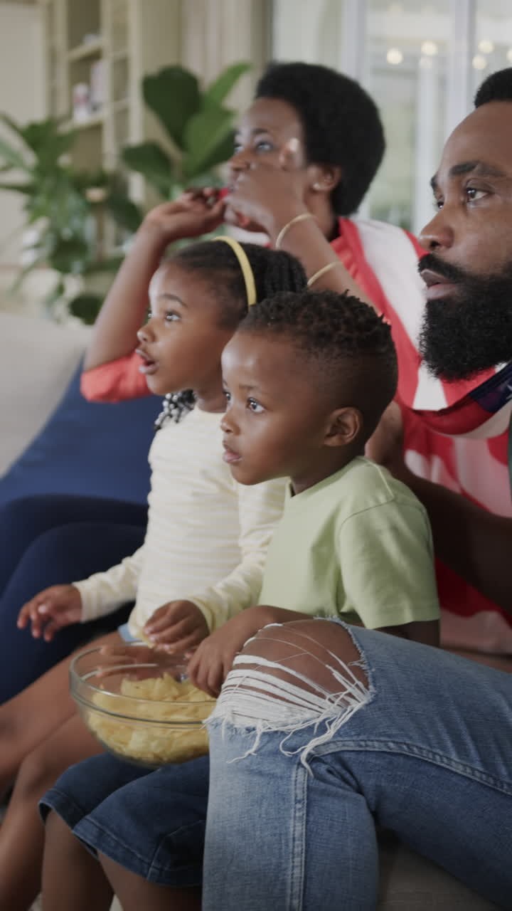 Vertical video of african american couple with son and daughter watching sport at home, slow motion