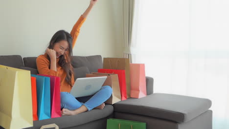 modelo asiático sentado en un sofá con las piernas cruzadas bolsas de compras en ambos lados, escribiendo en la computadora y riendo a carcajadas sonriendo