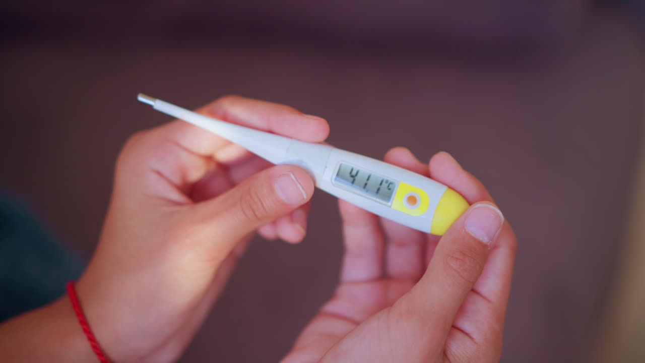 Close up of hands holding digital thermometer displaying high temperature of 41.1 degrees Celsius indicating fever or health emergency with focus on reading
