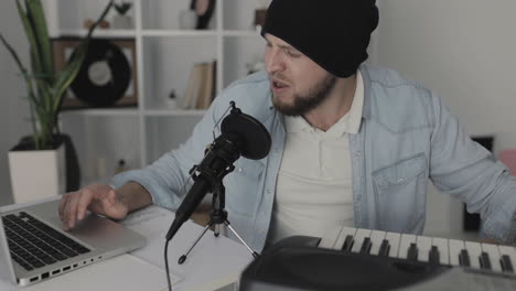 músico masculino cantando, tocando teclados eléctricos y usando una laptop en casa