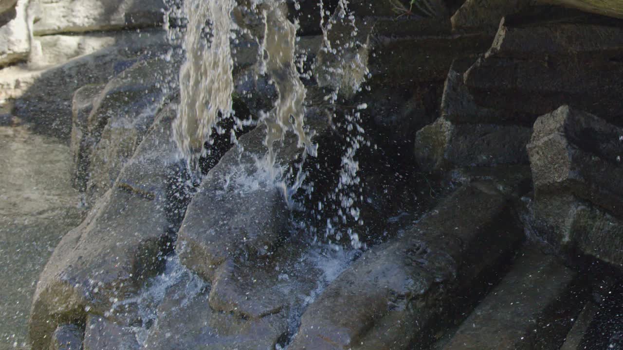 Cascading water flows over tiered stone fountain steps outdoors in bright natural daylight, close-up