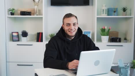 A-portrait-of-a-young-man-sitting-with-a-laptop-in-the-office,-smiling-and-looking-at-the-camera