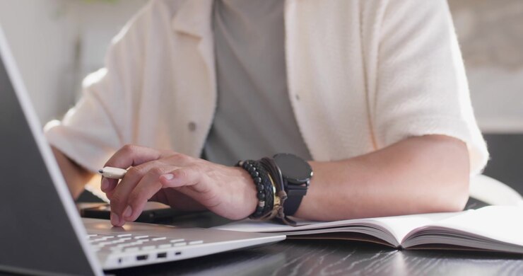 African American man typing on laptop and adjusting pen while checking notebook at office for work