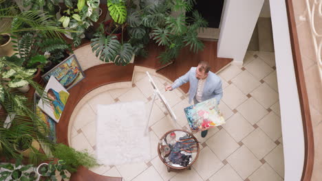 artist applies paint on canvas fixed on easel in plant gallery. professional painter holds palette drawing picture in home workshop. creative man improves talent