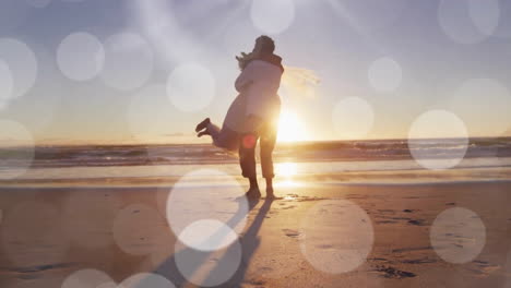 Animation-Von-Lichtpunkten-über-Einem-Glücklichen-Afroamerikanischen-Bräutigam,-Der-Die-Braut-Bei-Einer-Hochzeit-Am-Strand-Bei-Sonnenuntergang-Hochhebt