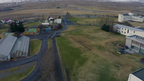 drone footage showcasing a small icelandic town featuring greenhouses, scattered buildings, open fields, and a snow-capped mountain in the background. a peaceful rural landscape.