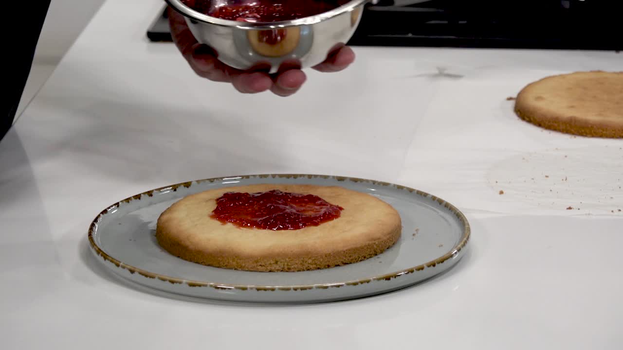 A person prepares a round cake by spreading fruit jam on top of a baked layer in a sleek kitchen. The process highlights the careful crafting of a delicious dessert