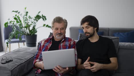 Young-man-teaching-old-dad-using-internet-online-with-computer-on-couch-in-living-room