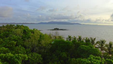 fly-over-a-little-tropical-wood-toward-a-little-rock-formation-in-the-sea-near-a-beach-where-some-people-are-hanging-out-during-a-warm-day