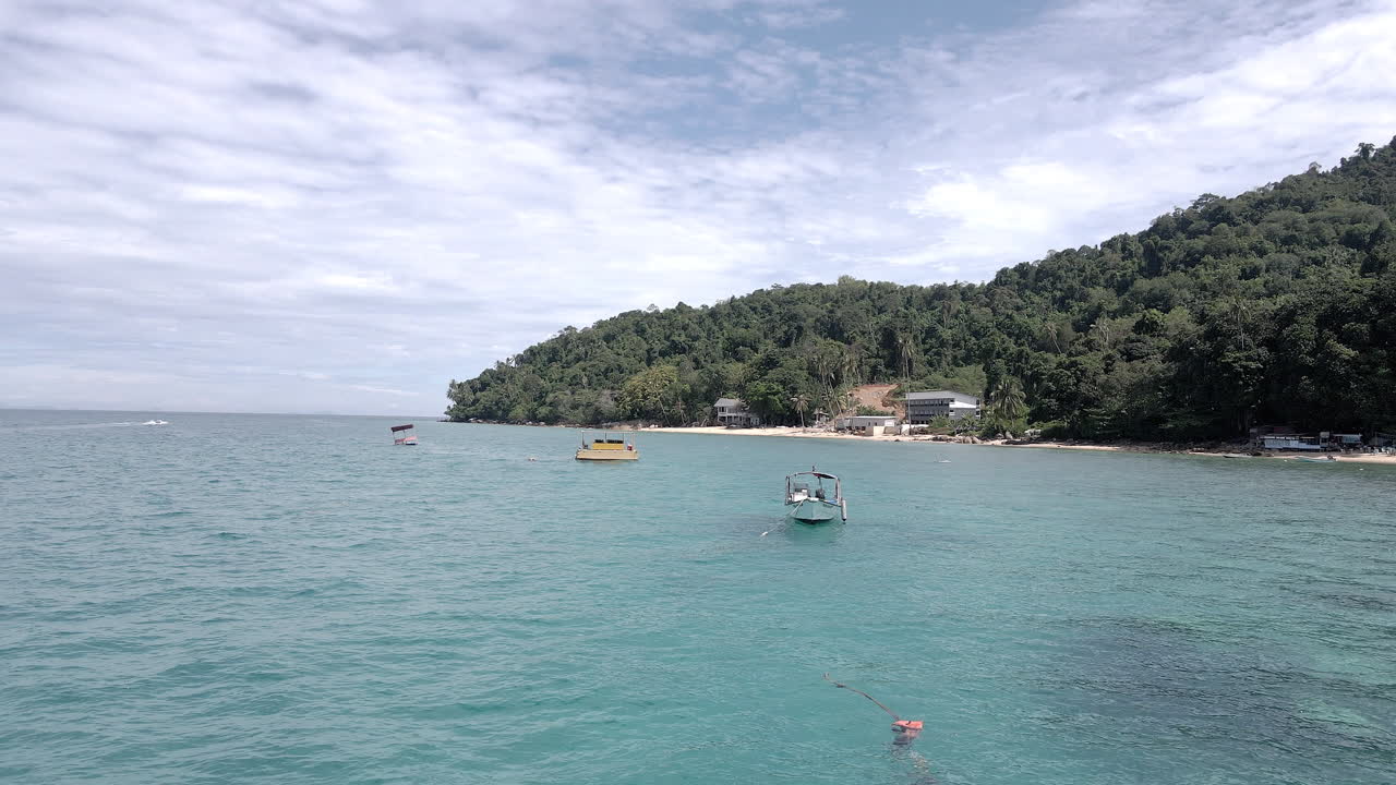 Aerial view of small boats floating on anchor in front of a remote island resort. Filmed at Alunan Resort, Perhentian Kecil, Malaysia. Rotating view above the ocean.