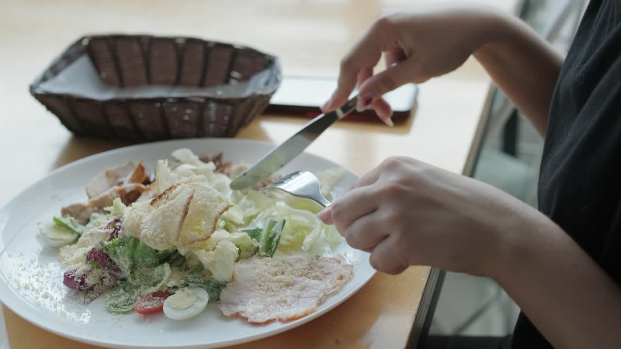 젊은 여성이 큰 창문, 자연빛, 도시 전망을 가진 현대적인 레스토랑에서 나무 테이블에서 신선한 러드를 먹고 있습니다. 편안한 식사 장면