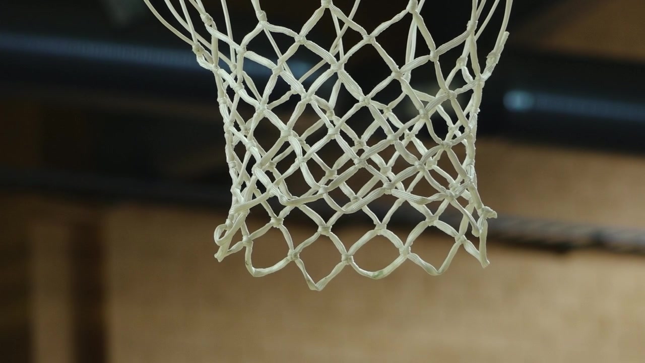 Primo piano di un pallone da basket che cade attraverso una rete bianca in un campo al coperto, con una leggera sfocatura di movimento e una messa a fuoco ridotta che enfatizzano l'azione e lo sport.