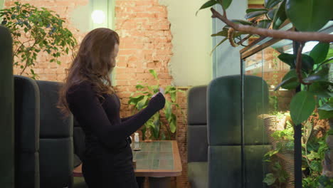 woman steps back with gentle smile while holding phone to photograph lush green environment enclosed in glass, surrounded by cozy booth seating and soft lighting in stylish brick-wall interior