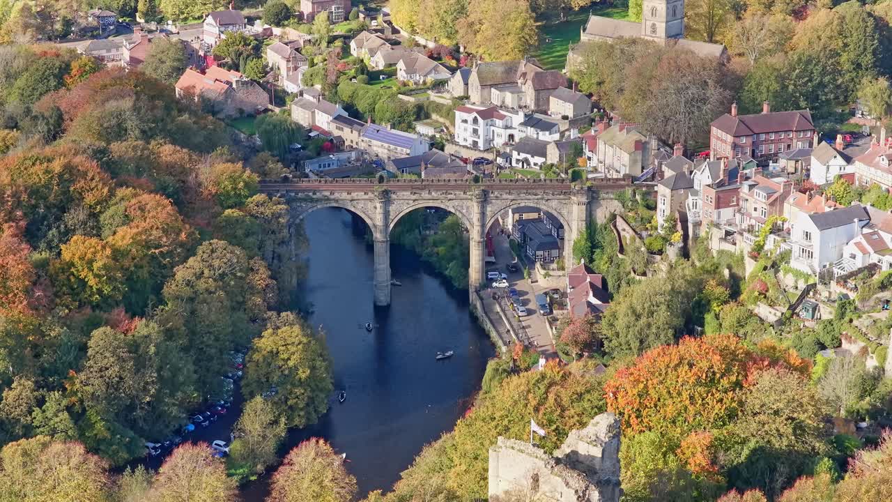 Aerial pull back from Knaresborough Viaduct reveals stone railway bridge above River Nidd, boats below, golden autumn woodland, urban streets, and medieval architecture in North Yorkshire, England