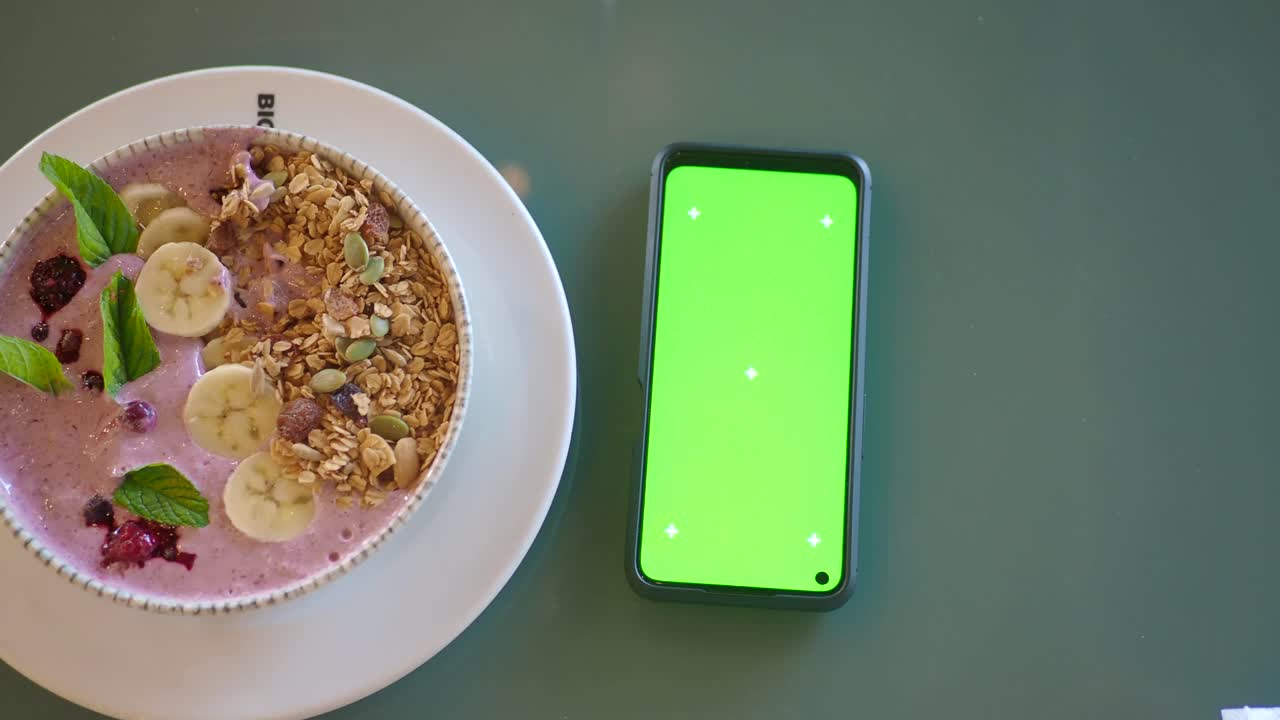 Smoothie bowl with granola and banana next to a smartphone with green screen