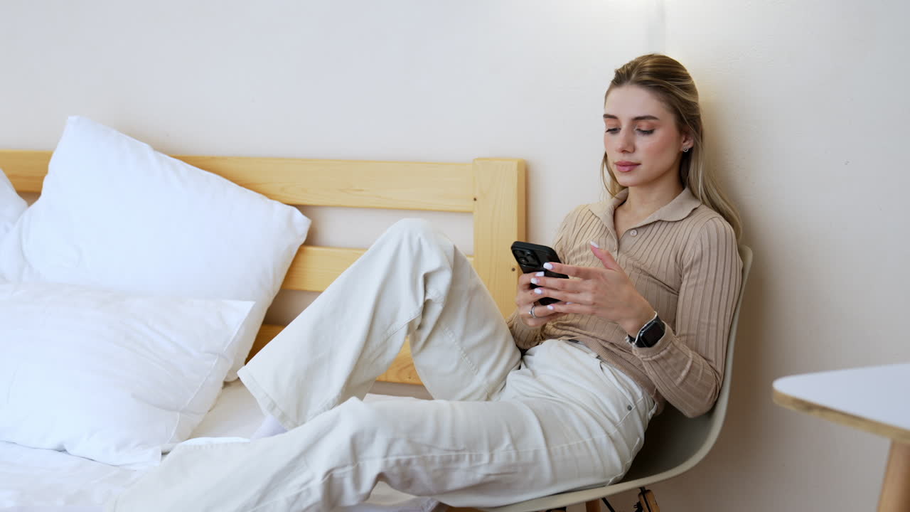 Blonde woman sitting on the chair resting her legs on the bed. Lady uses her smartphone.
