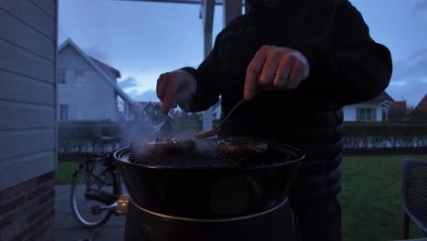 manos girando una hamburguesa en una parrilla con llamas, fuego y humo en un jardín durante el crepúsculo