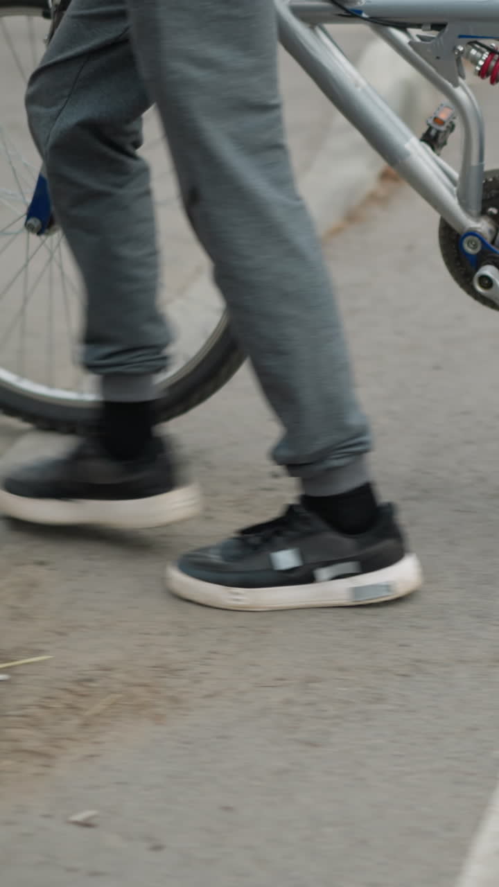 Leg view someone in grey pants and sneakers walking alongside a bicycle across a pedestrian crossing, with part of a car approaching, and poles with red markings by the roadside
