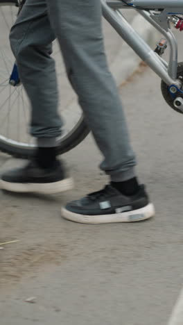 leg view someone in grey pants and sneakers walking alongside a bicycle across a pedestrian crossing, with part of a car approaching, and poles with red markings by the roadside