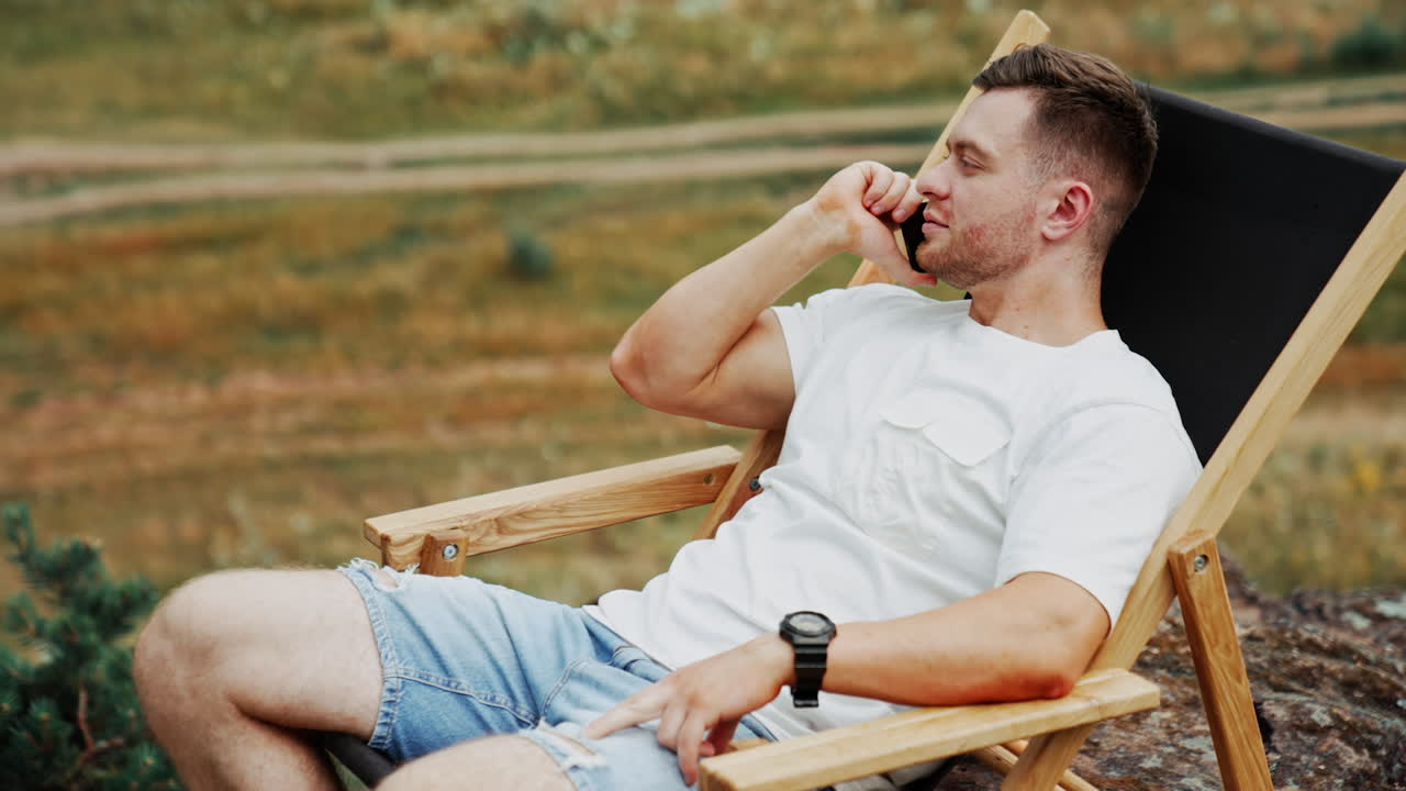 Calm relaxed man wearing white t-shirt and jeans shorts outdoors. Man speaks on the phone sitting in a comfortable folding chair.